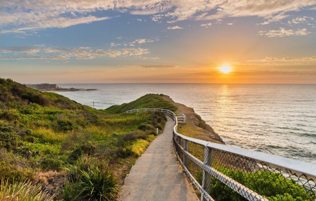 Walking path with white railing leading toward the ocean at sunrise, with lush greenery and golden sunlight reflecting on the water.