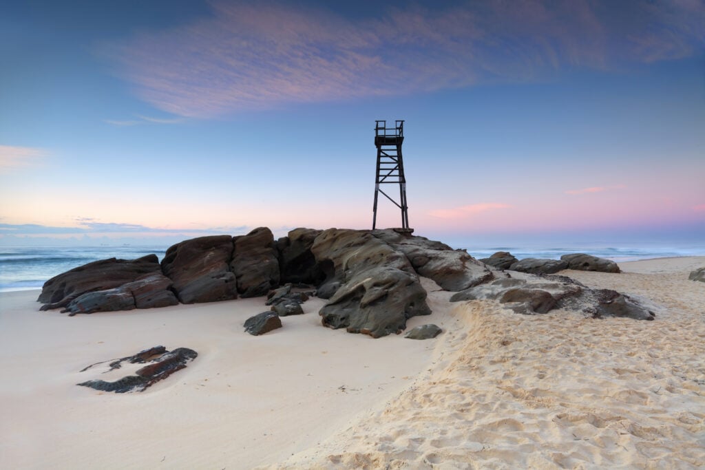 A tall wooden lifeguard tower stands on rocky formations by a sandy beach at sunrise, with pastel colours in the sky.