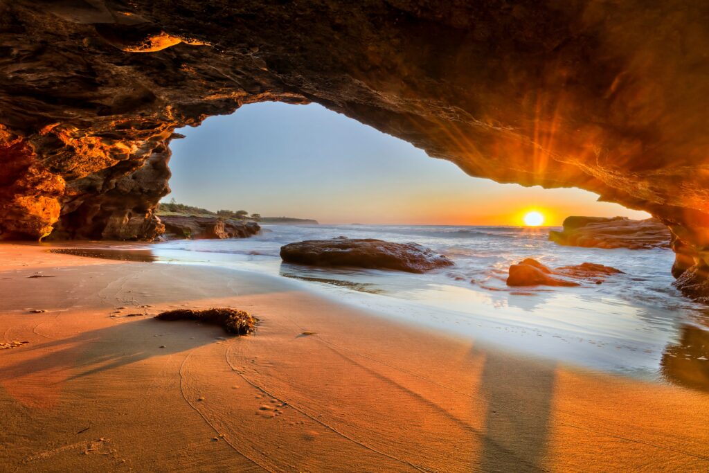 Sunrise view from inside a rocky coastal cave in Newcastle, with sunlight streaming across the sandy beach and waves crashing outside.