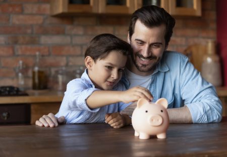 Shartru Wealth father teaching son about saving money by putting coins in a piggy bank together.