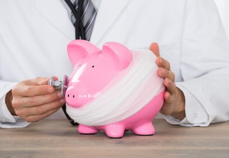 A medical professional listens to a piggy bank with a stethoscope featured in Shartru Wealth article.