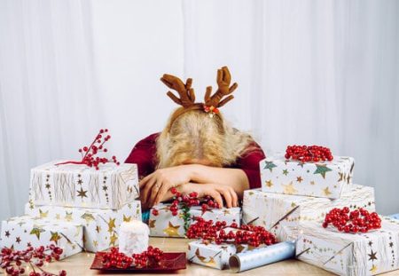 Exhausted woman with reindeer antlers surrounded by Christmas gifts, budgeting stress explained by Shartru Wealth Management.