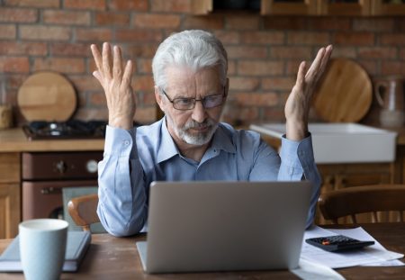 Shartru Wealth image of frustrated older man raising hands while looking at laptop with bills.