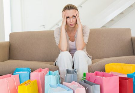 A Shartru Wealth photo shows a frustrated woman sitting on a couch with shopping bags.