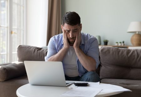 Shartru Wealth image of stressed man at home holding head while checking bills on laptop.
