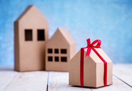 A photo from Shartru Wealth shows a small cardboard house with a red ribbon tied around it.