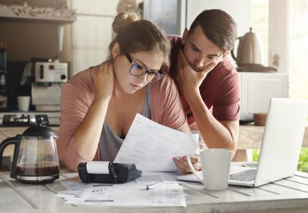 Couple reviewing bills at home worried about household debt comparison by Shartru Wealth.