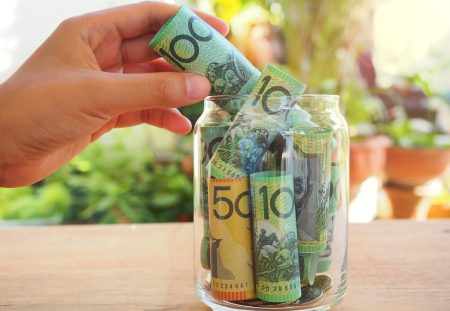 Shartru Wealth hand placing Australian banknotes into a glass savings jar.