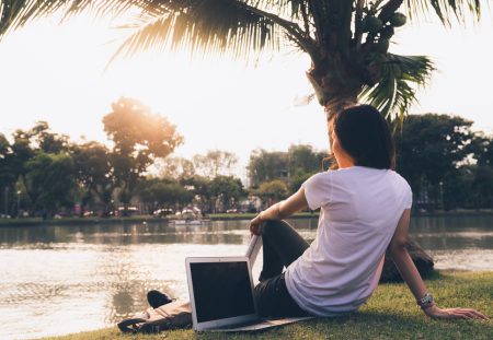 A person from Shartru Wealth works on a laptop under a palm tree beside a lake.