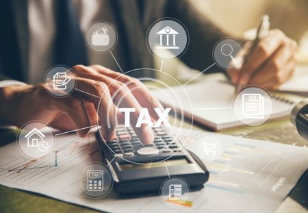 A person's hands press on a calculator showing the word "TAX" for Shartru Wealth.