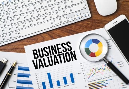 Business valuation document with graphs, calculator, pen, smartphone, and computer keyboard on desk.