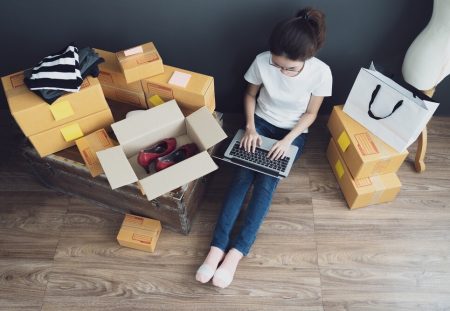 Young woman selling unwanted items online with parcels, laptop, and boxes, Shartru Wealth Management.