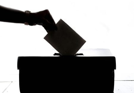 Hand placing a ballot into a ballot box, used in a Shartru Wealth article.